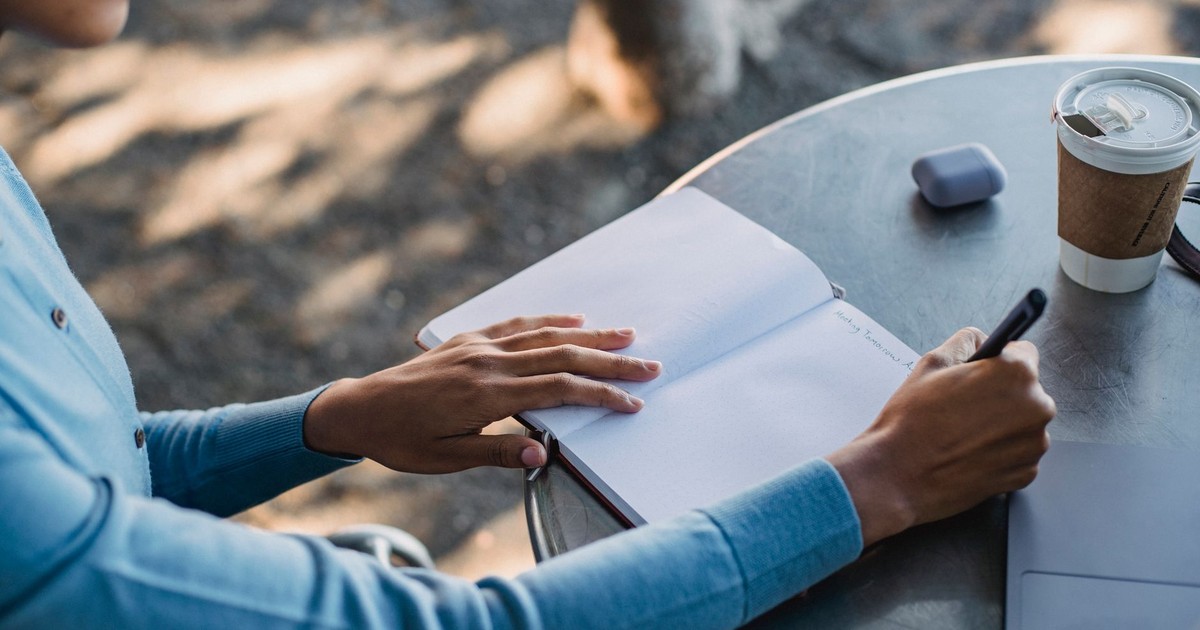A close-up of a person writing notes in a notebook at an outdoor cafe table with a coffee cup.