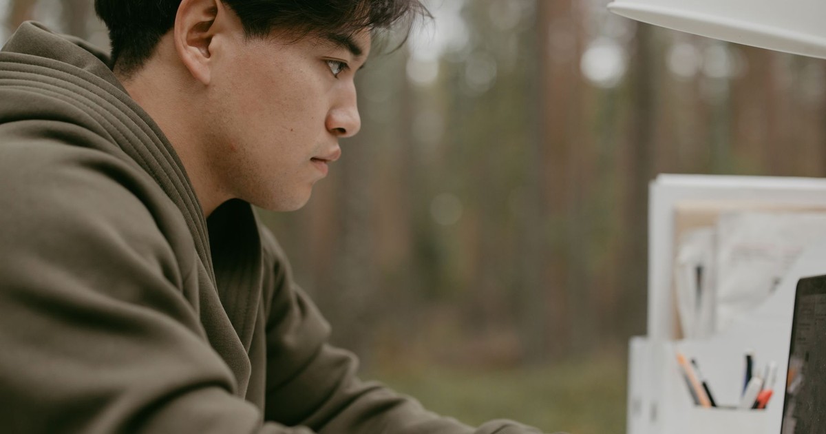 Young man in a green jacket using a laptop outdoors, concentrating on his work.