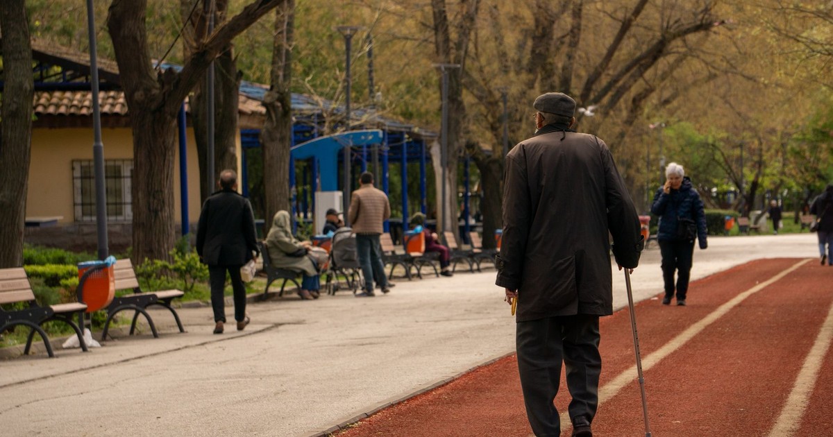 An elderly man with a cane strolls down a serene park path lined with benches and trees.