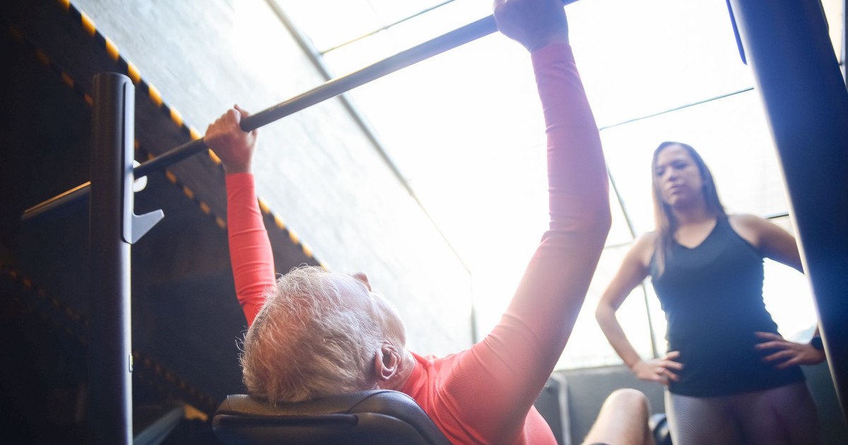 Senior man bench pressing with a female trainer overseeing his workout in an indoor gym.