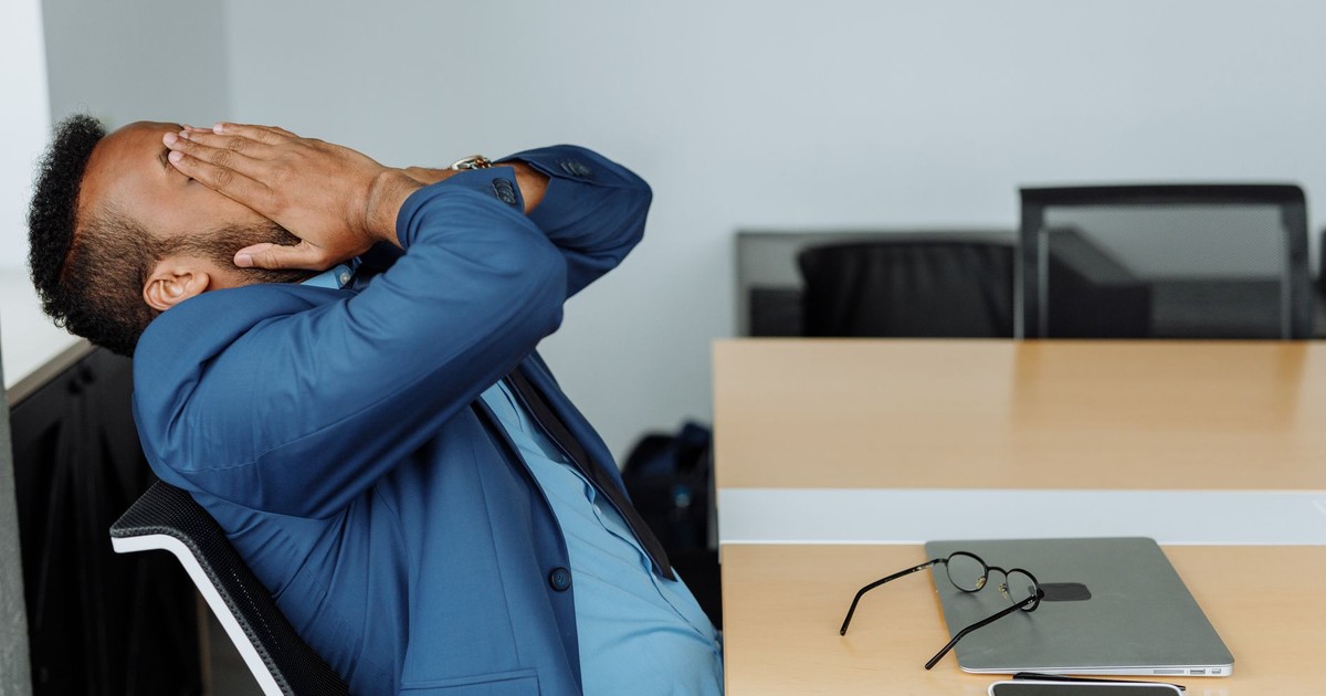 A stressed businessman in a suit covers his face while sitting at a desk with office tools.
