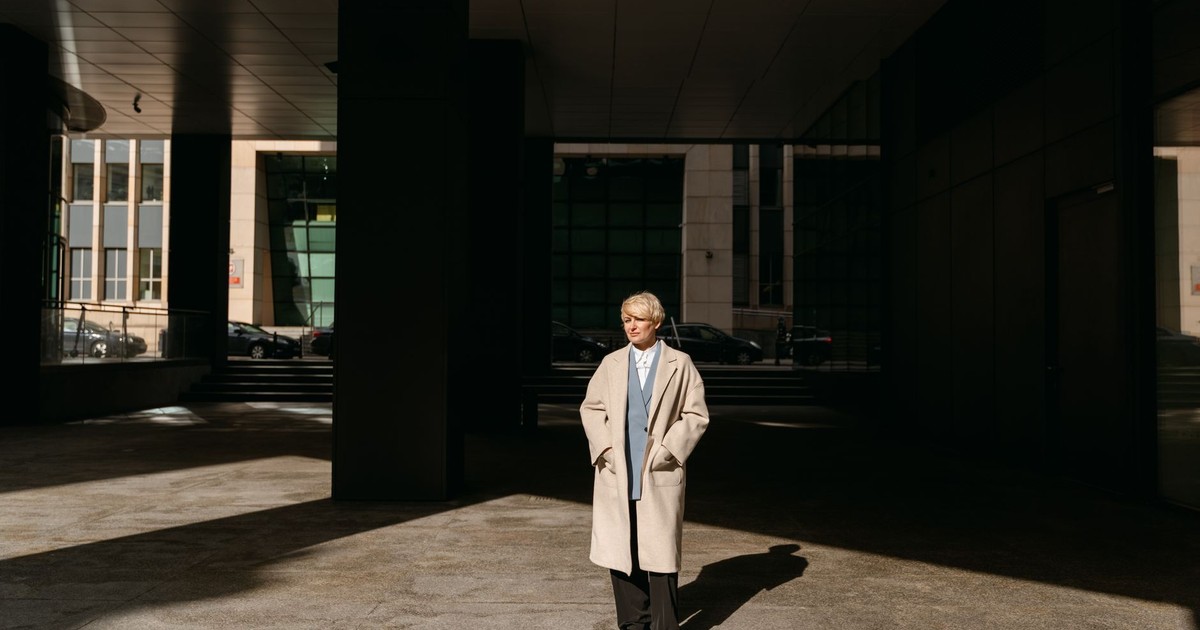 A woman in a coat stands confidently in an urban shadowed area, hands in pockets, modern style.