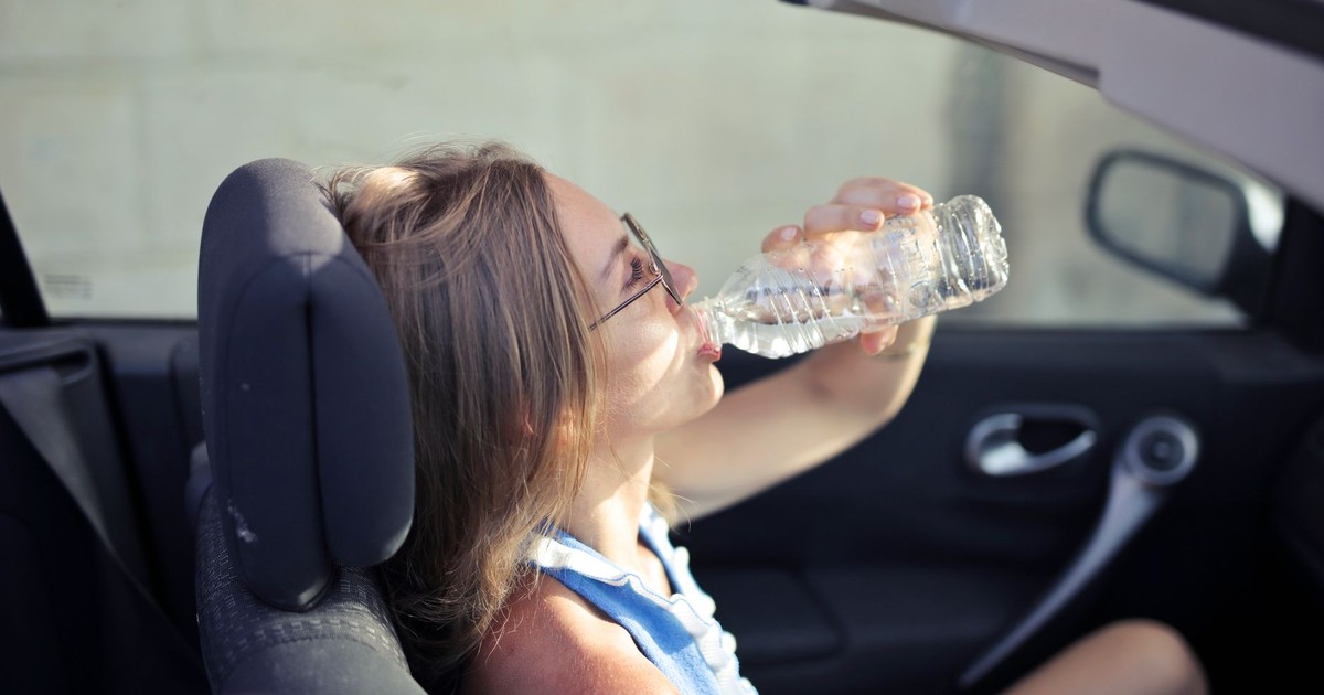 High angle side view of young woman in glasses and casual clothes drinking water from plastic transparent bottle while sitting in cabriolet with open roof in traffic jam in hot sunny day