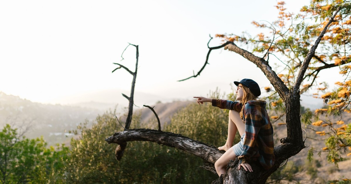 A young girl sits on a tree trunk outdoors, pointing towards a scenic view.