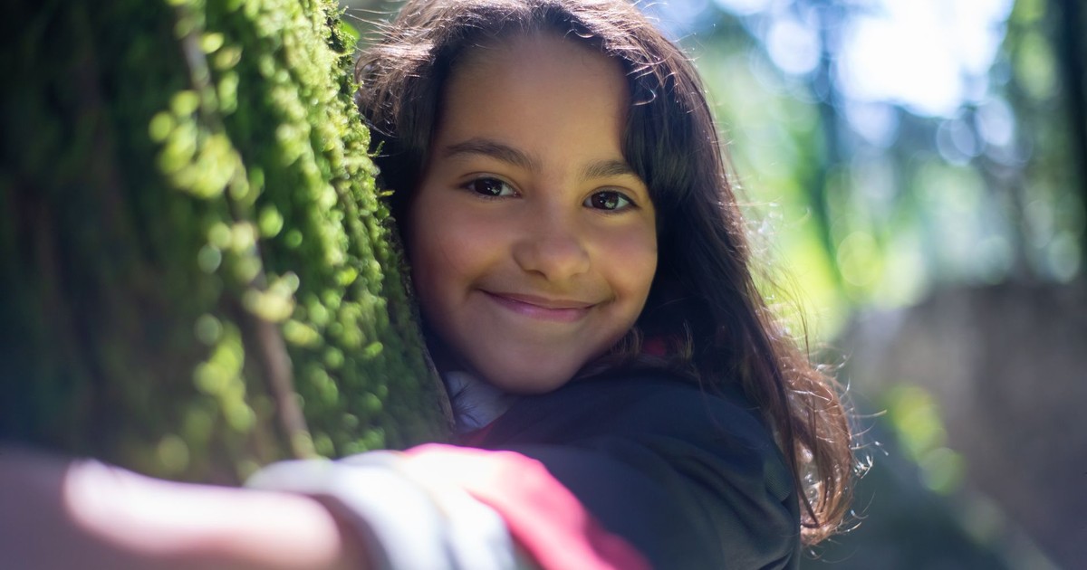 A cheerful young girl hugs a moss-covered tree trunk, enjoying the natural surroundings.