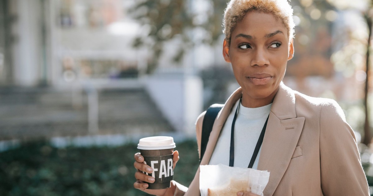 Content young African American female entrepreneur drinking coffee to go and eating sandwich while going to appointment in park and looking away