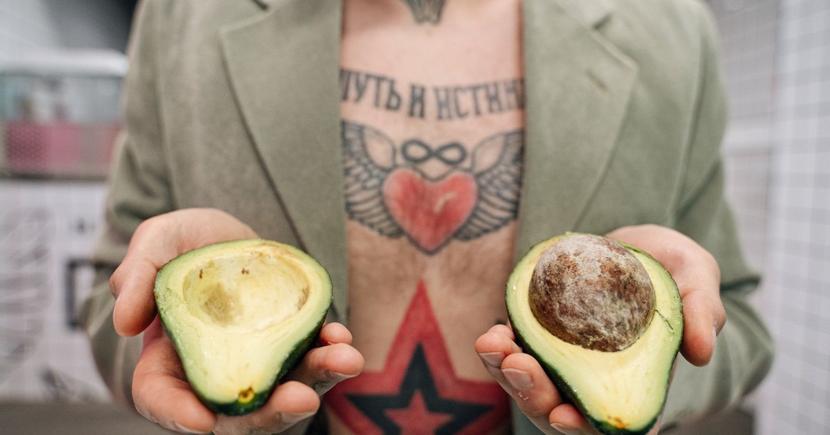 A tattooed man in a jacket presents two halves of an avocado indoors, showcasing unique chest art.