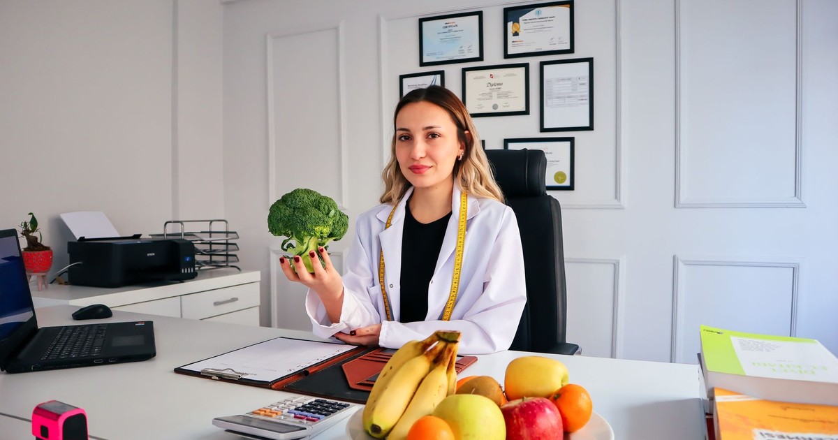 Female nutritionist in office holding broccoli, surrounded by fruits, promoting healthy lifestyle.