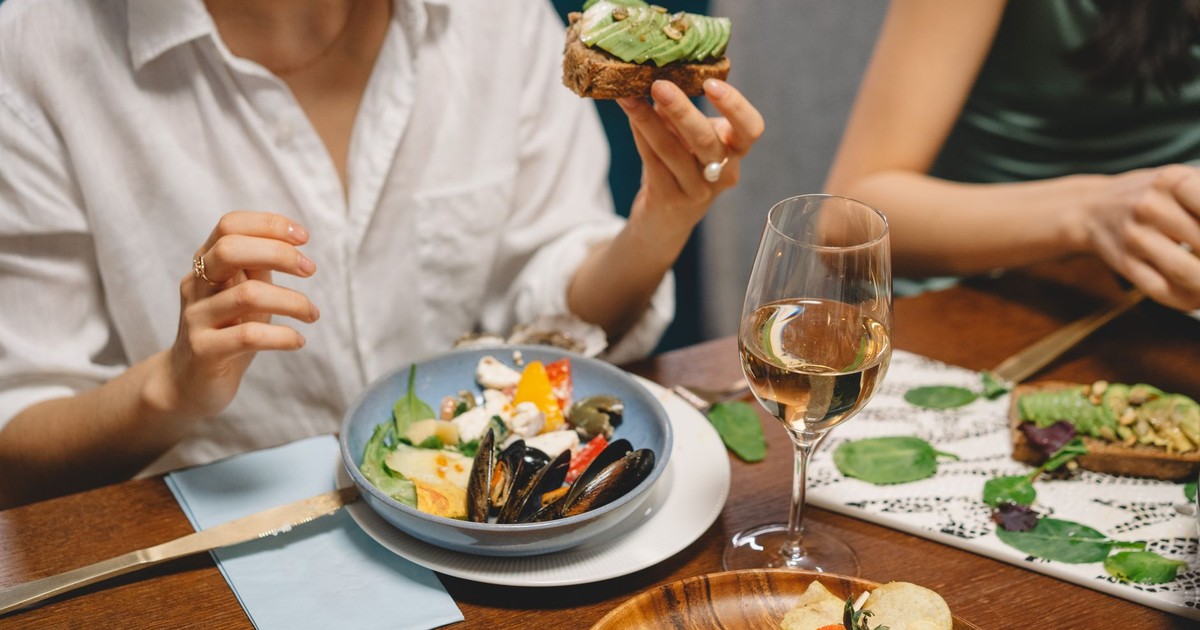A colorful dining scene featuring avocado toast, seafood, and white wine shared among friends.