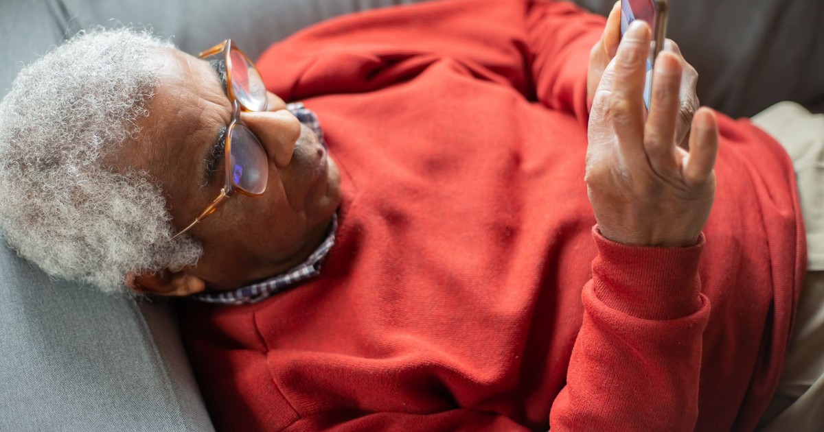 Senior man in red sweater using smartphone while lying on a sofa, enjoying leisure time indoors.