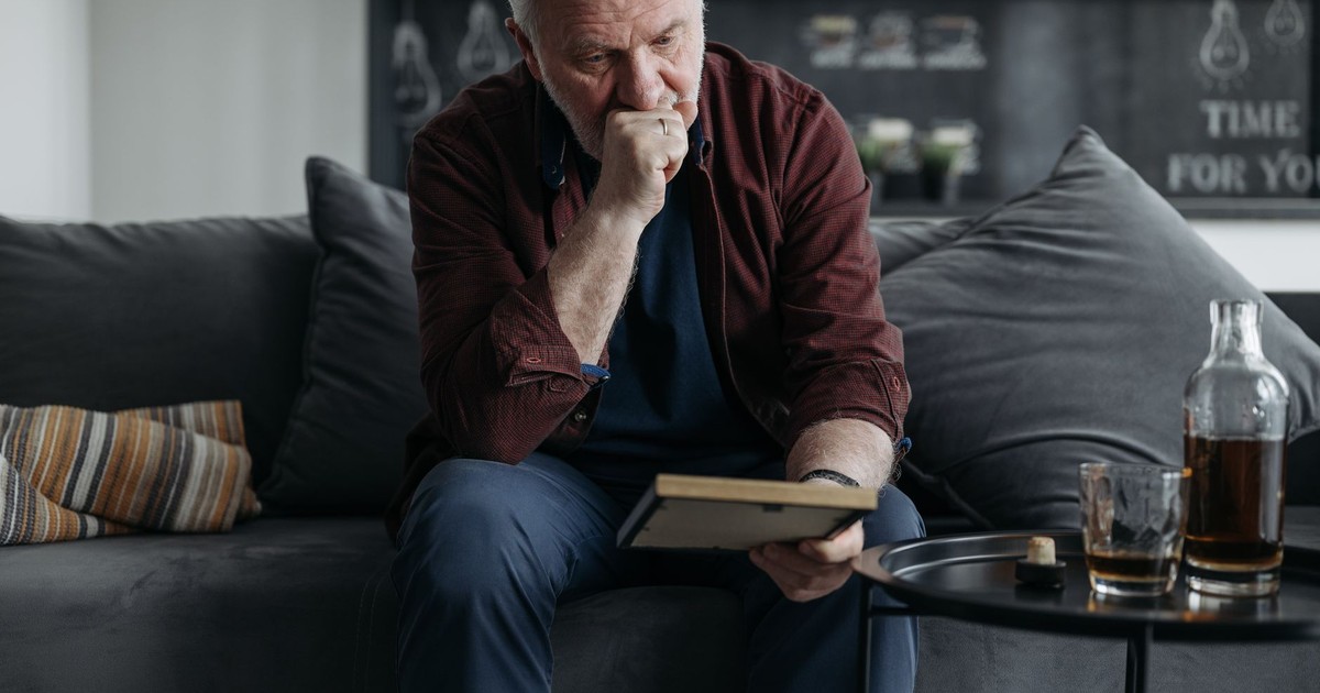 A senior adult man sitting on a couch, lost in thought while reading a book indoors.