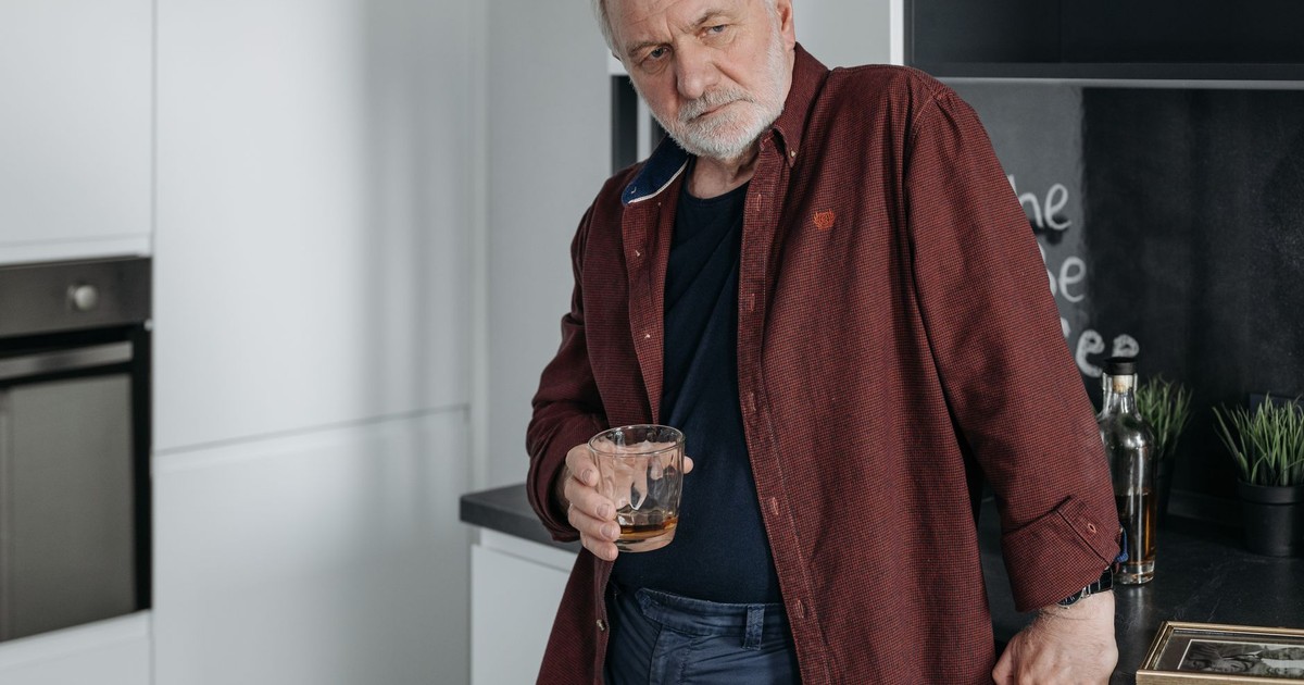 Senior man in a modern kitchen holding a drink, expressing deep emotion and contemplation.