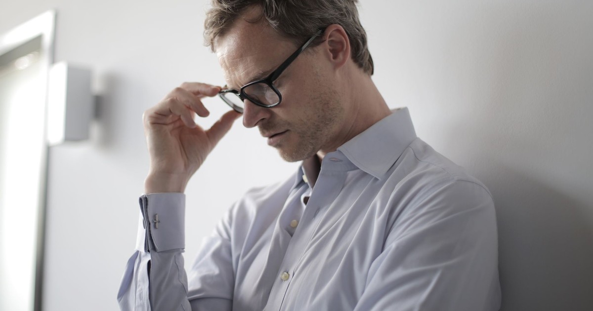 An adult man in a shirt and glasses appears stressed while leaning against a wall indoors.