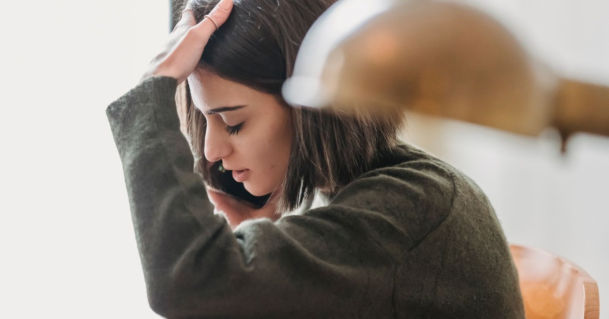 Side view of upset young ethnic female millennial with dark hair grabbing head with closed eyes while having phone conversation sitting on chair at home