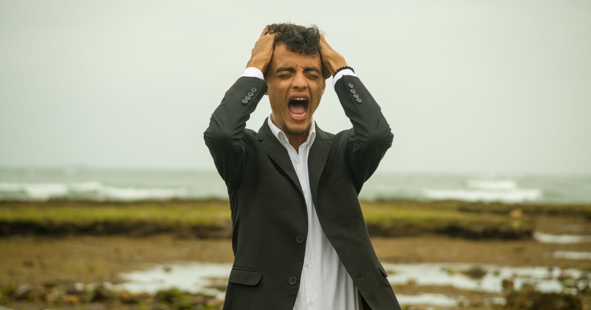 A man in a suit screams in frustration while standing on a windy beach. Emotional stress concept.