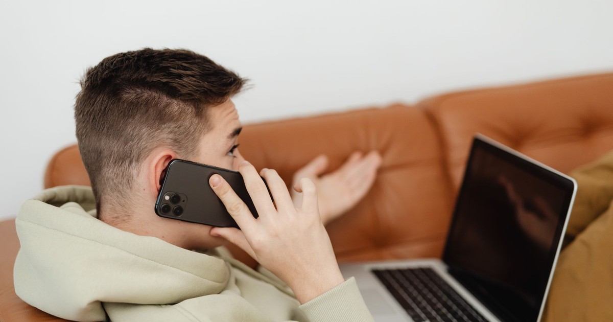 A young man in a hoodie talks on his phone while using a laptop on a brown couch.