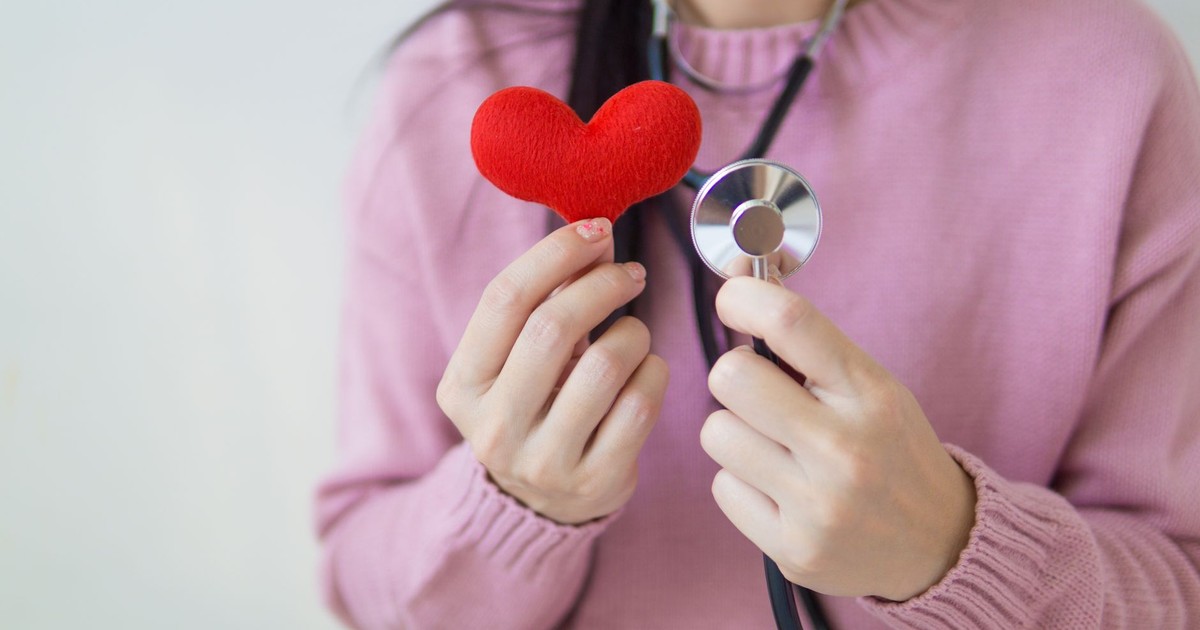 Unrecognizable female in pink sweater with stethoscope on neck standing on white background with red heart in hand in daylight