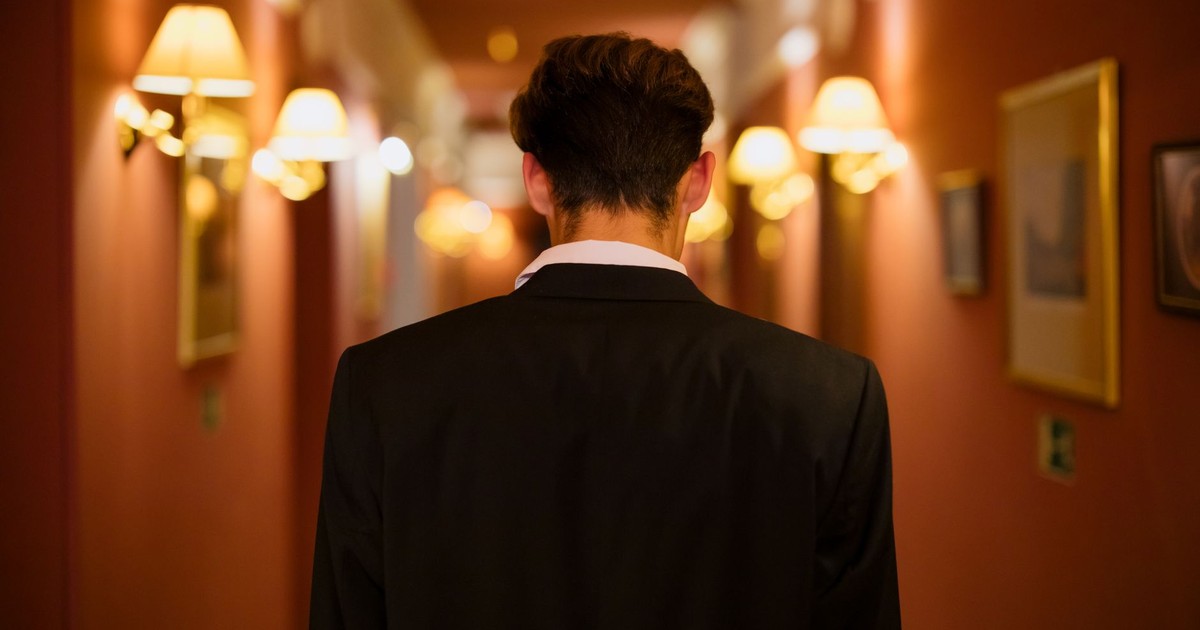Rear view of man in black suit walking down a warmly lit hotel corridor.