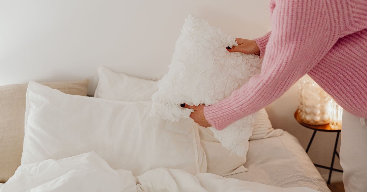 A person in a pink sweater arranges pillows in a cozy, well-lit bedroom.