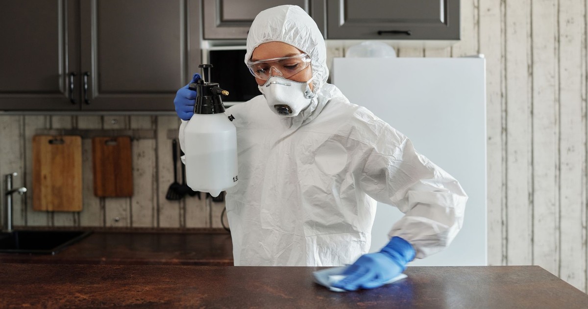A person in protective clothing disinfects a kitchen counter indoors, focusing on safety and cleanliness.