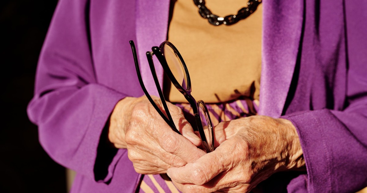 An elderly woman in purple clothing holds eyeglasses, highlighting age and elegance.