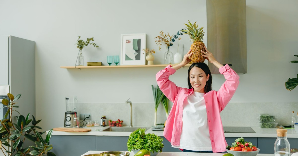 Asian woman in pink overshirt holding a pineapple in a modern kitchen with fresh produce and cheerful ambiance.
