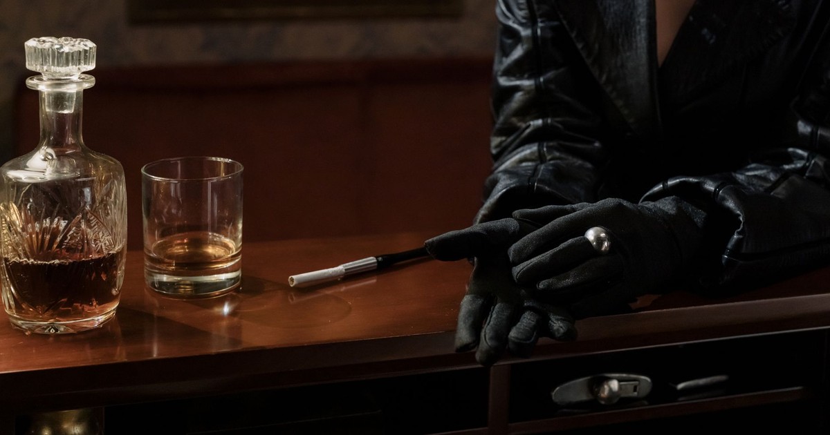 Dark and cinematic scene with glass of whiskey, cigarette, and black gloves evoking a noir atmosphere.