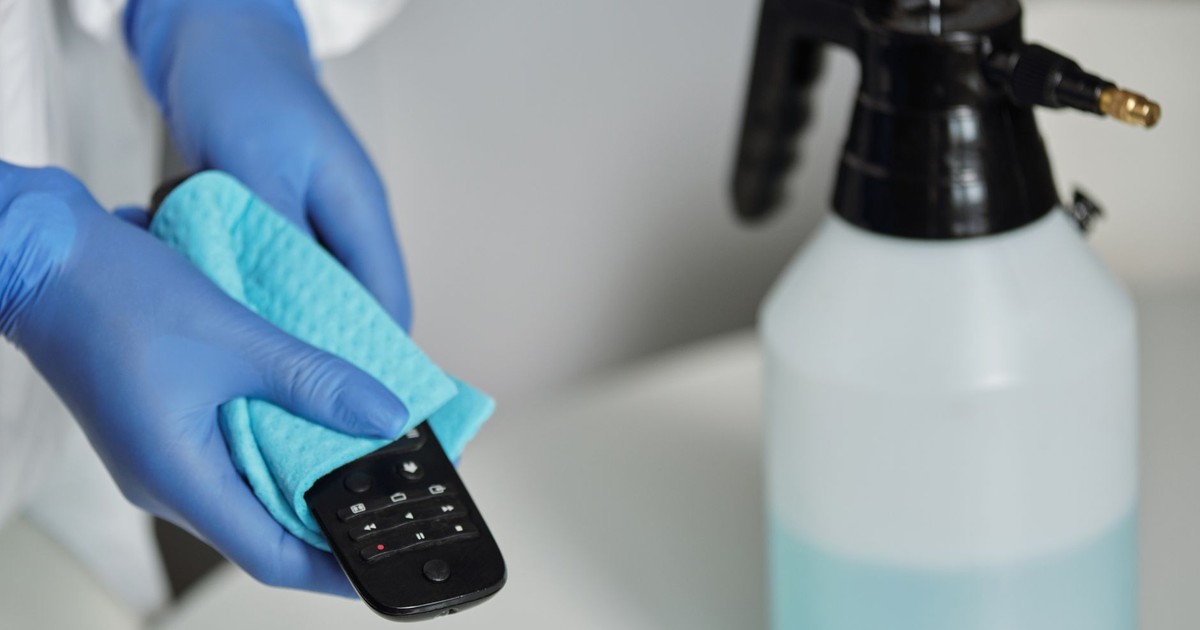 Close-up of gloved hands disinfecting a remote control with a cloth and spray bottle.