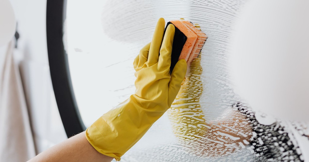 Anonymous person in yellow rubber gloves using simple sponge for cleaning mirror in bathroom