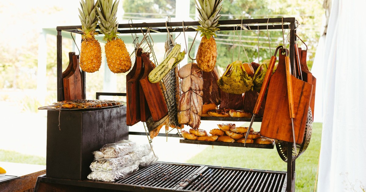 A rustic outdoor barbecue with pineapples, vegetables, and meat grilling under the sun.