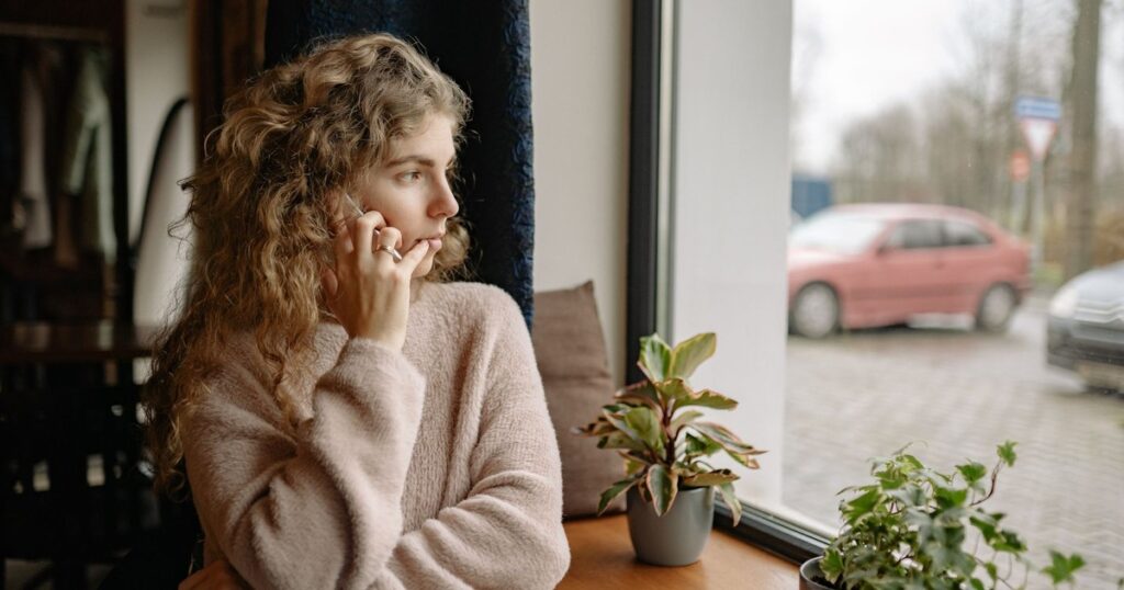 Woman in cozy sweater talking on a phone by the window, surrounded by houseplants, observing outdoors.