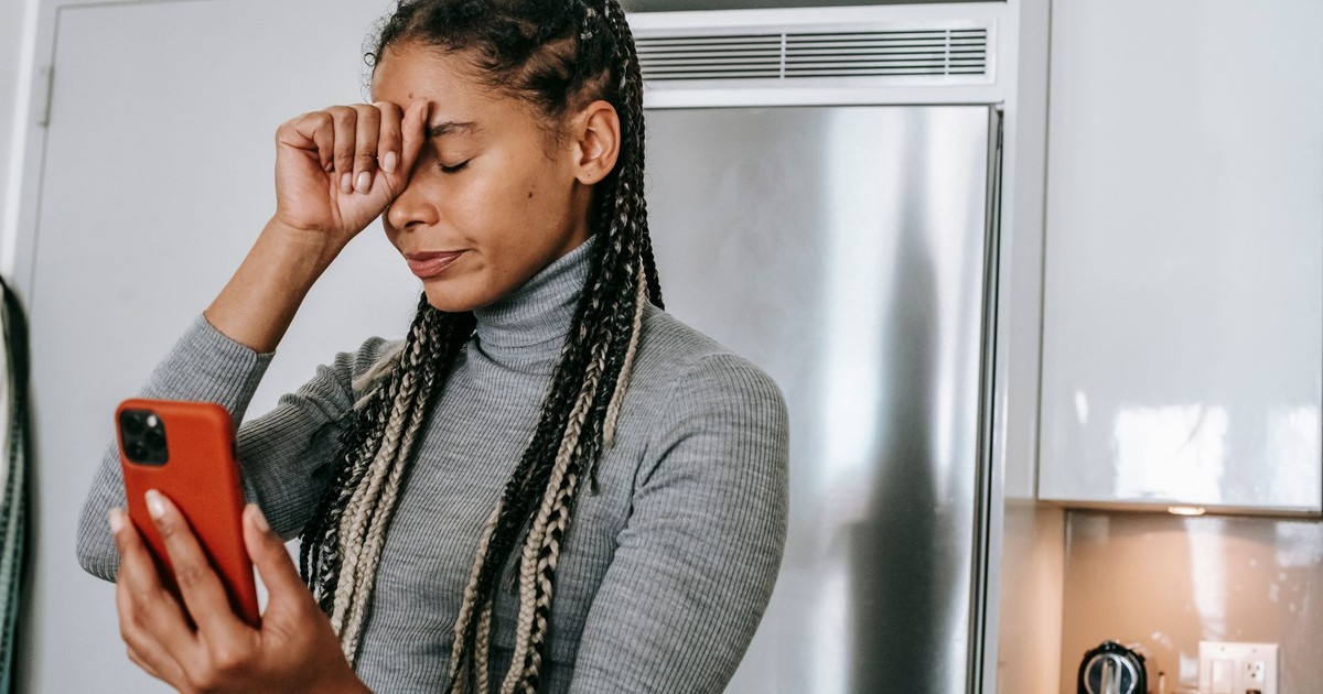 Unhappy young African American female with long braids in turtleneck touching face with closed eyes while having video conversation on smartphone in kitchen