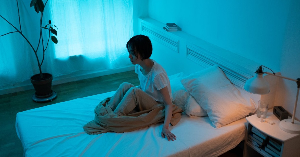 Woman sitting on bed in dimly lit room, conveying themes of insomnia and contemplation.