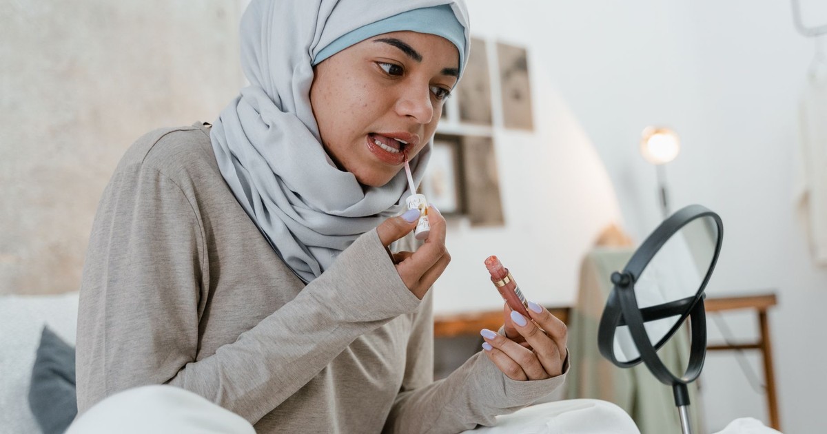 Young Muslim woman applying lipstick while sitting in a cozy bedroom setting with mirror.