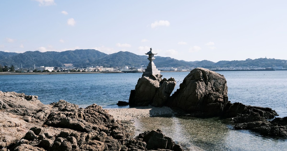 Tranquil coastal view of Gamagōri, Japan, featuring a stone lantern on rocky shorelines with scenic mountain backdrop.