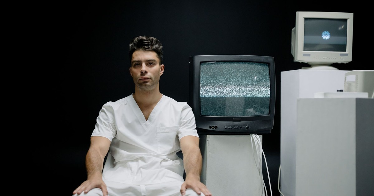 A serious man in white attire sits beside vintage TVs with static screens.