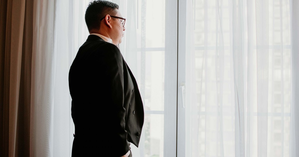 A man in a suit gazes pensively out a bright window indoors.