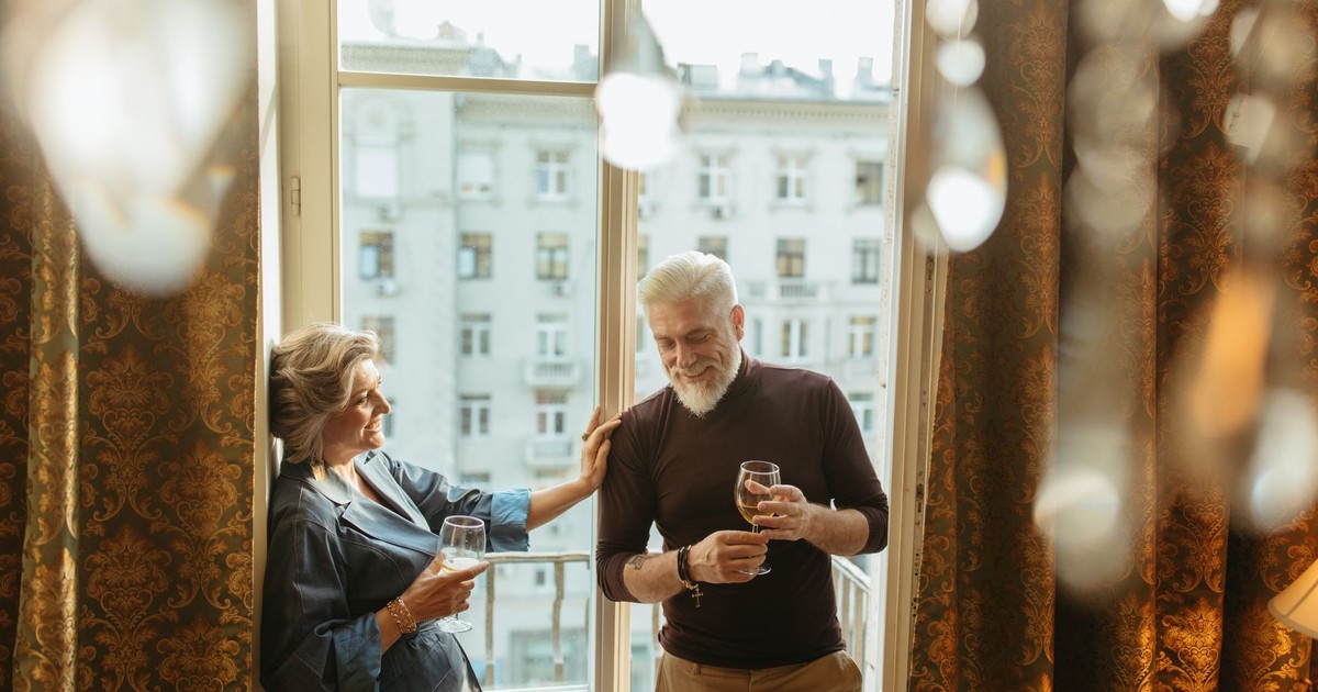 A senior couple enjoys wine and laughter by balcony windows inside a cozy apartment.