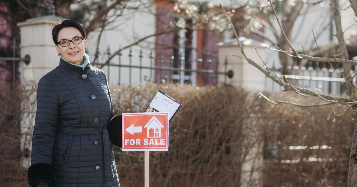Smiling woman realtor in winter coat standing by a for sale sign with documents, outdoors.