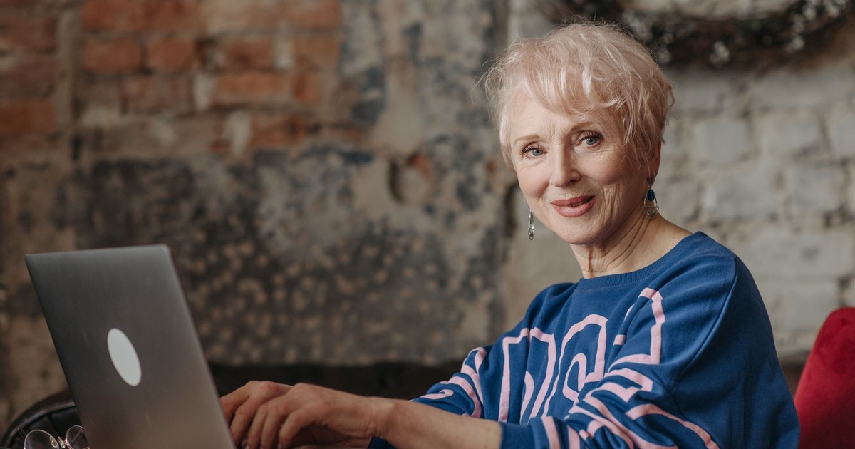 Elderly woman using a laptop at home office with rustic brick wall. A glimpse into modern senior lifestyle.