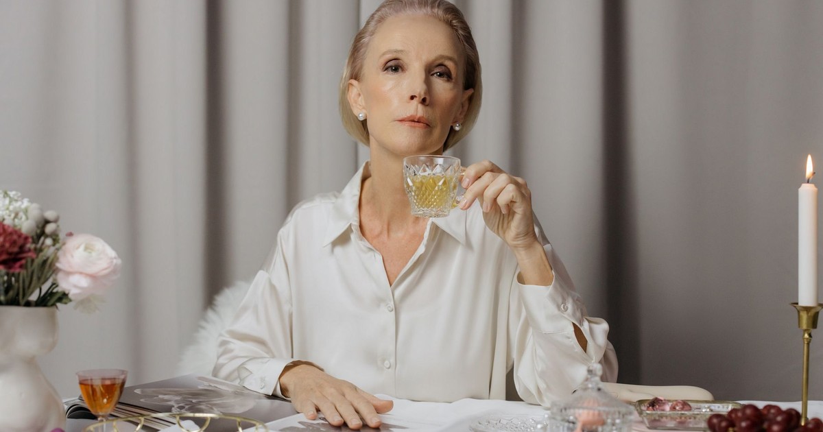Senior woman in white blouse sipping a drink in an elegant setting with flowers and candlelight.