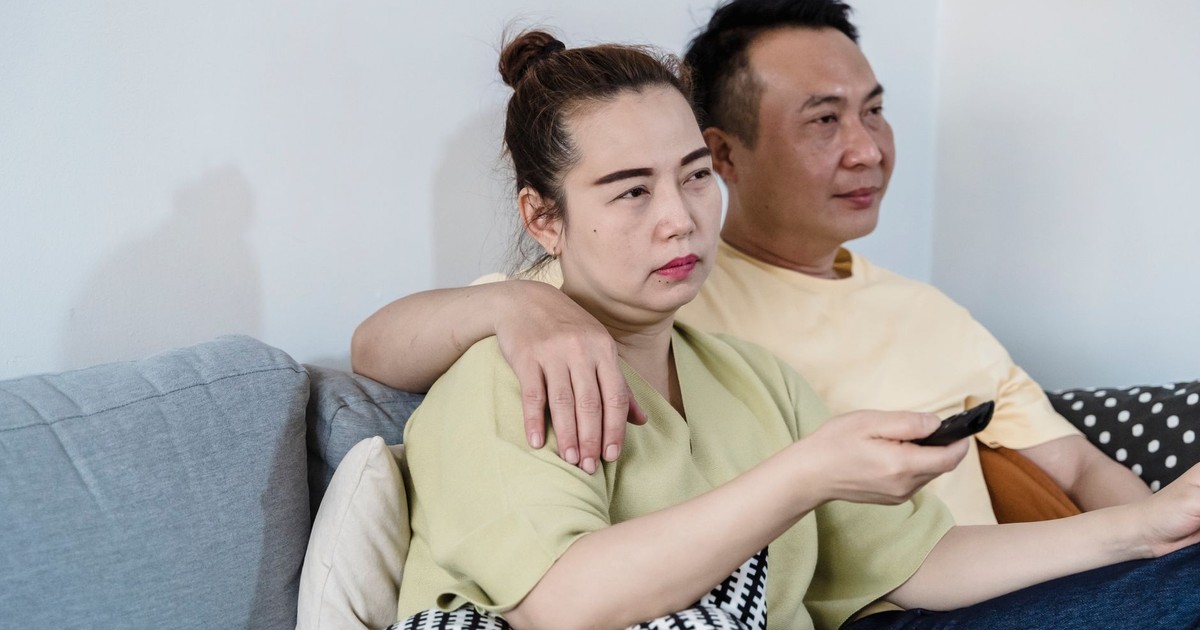 An Asian couple sitting on a couch at home, watching TV together with a remote control in hand.