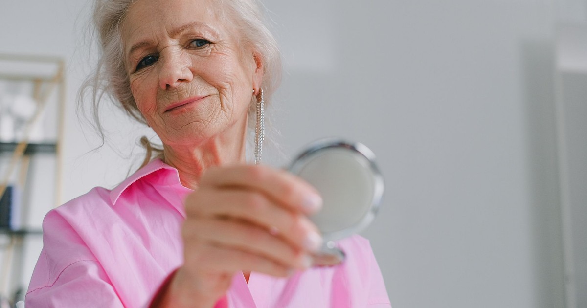 Elegant elderly woman in pink shirt holding a compact mirror indoors, reflecting joy and confidence.