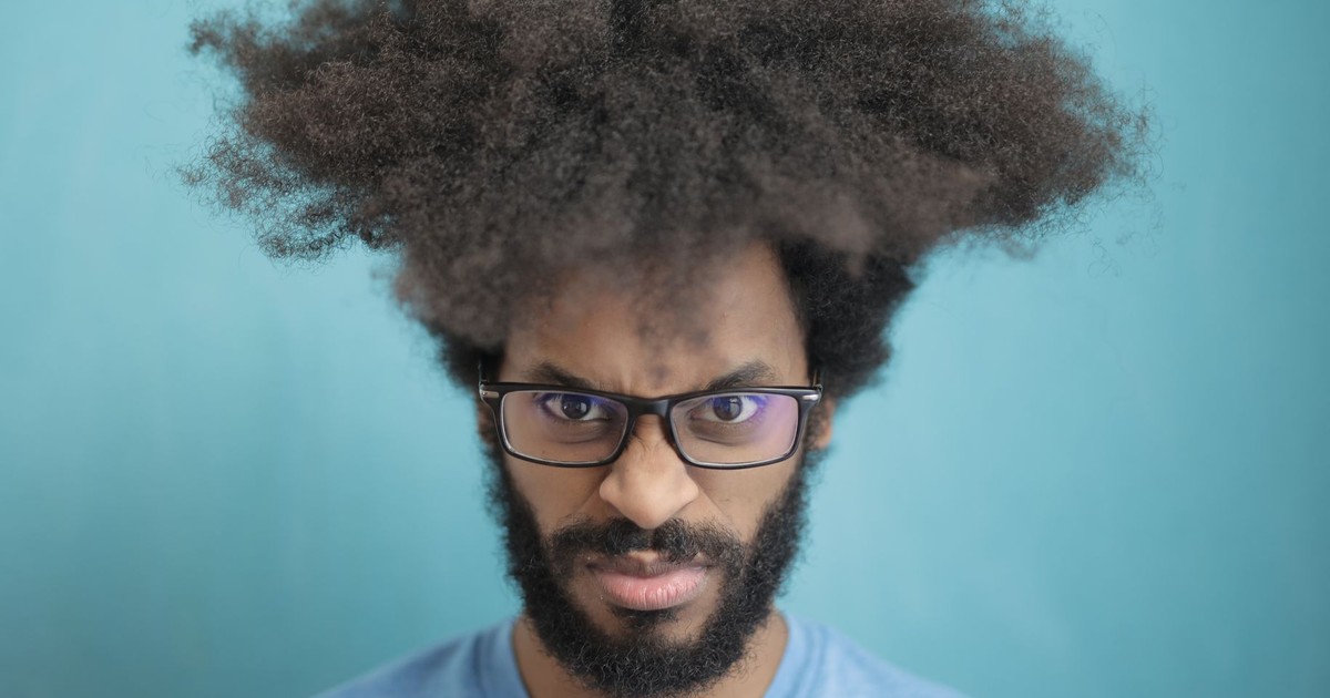A man with afro hairstyle and beard looking intense, wearing glasses, on a blue backdrop.
