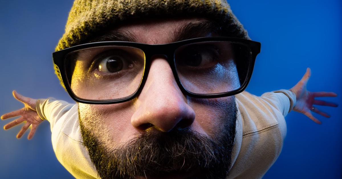 Close-up fisheye portrait of a bearded man wearing glasses and a beanie against a blue background.