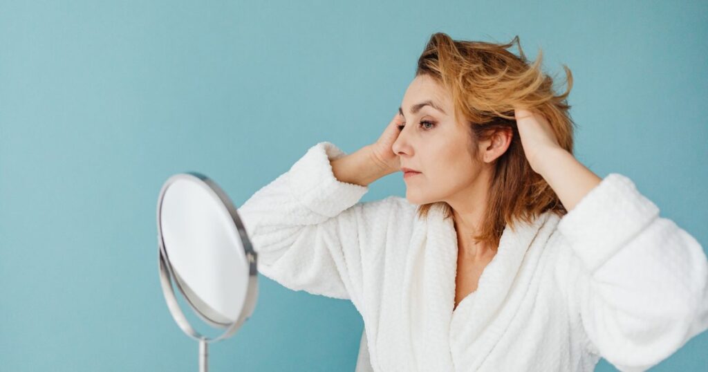 Adult woman in a bathrobe fixing her hair in front of a mirror against a blue background.