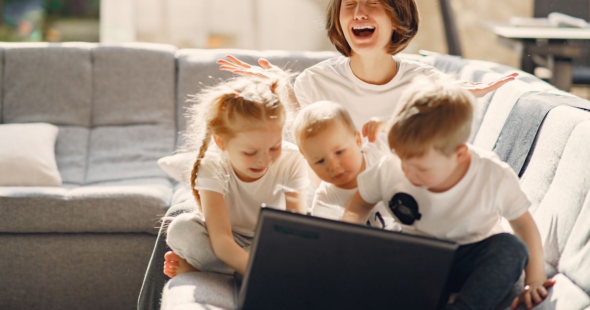 Stressed woman asking for help while sitting with small children playing on laptop together on sofa in living room