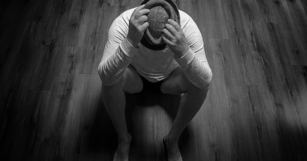 Black and white photo of a person sitting with a hat on a wooden floor, conveying sadness.
