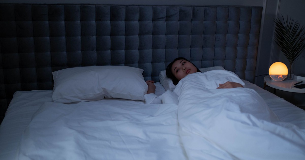 An Asian woman lying awake under soft white bedding in a dimly lit modern bedroom.