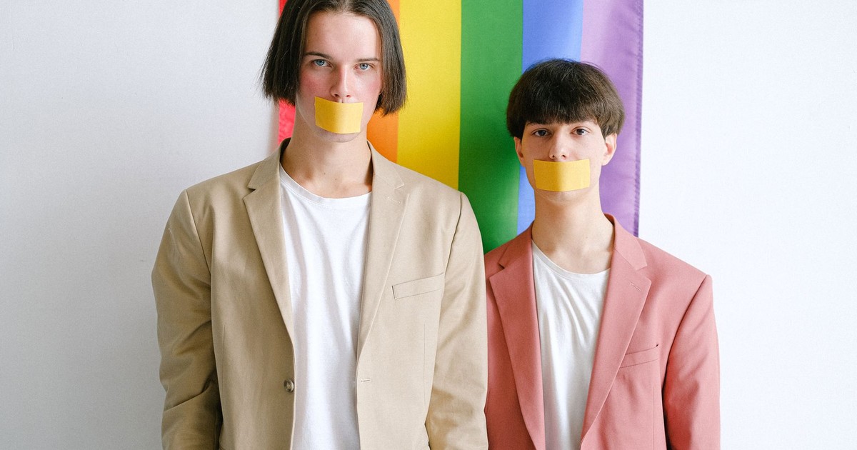 Two men with taped mouths stand against a rainbow flag, symbolizing LGBTQ silence and oppression.